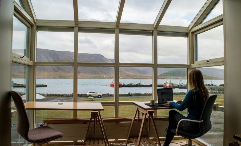 Person in a blue jacket working on a laptop at a wooden desk by a window