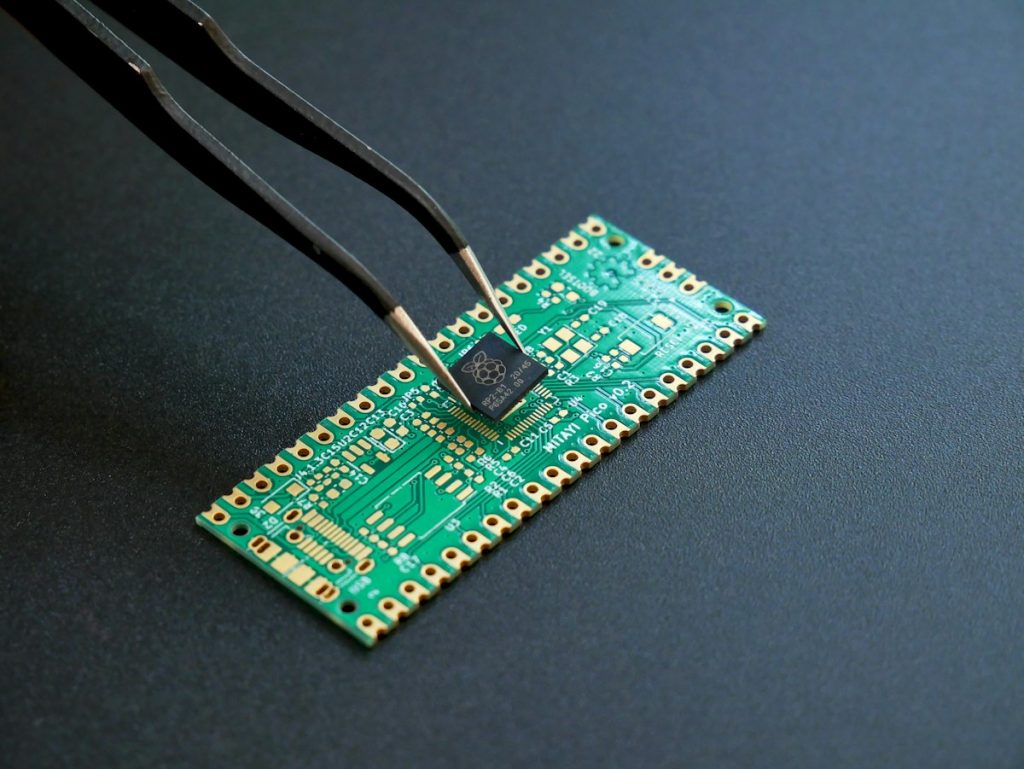 Tweezers holding a small packaged microcontroller chip above a green printed circuit board